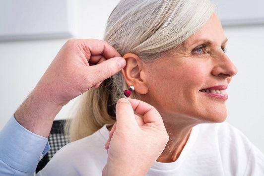 Ältere Frau erhält Hörgerät, Hörakustiker passt modernes Hörgerät am Ohr an.